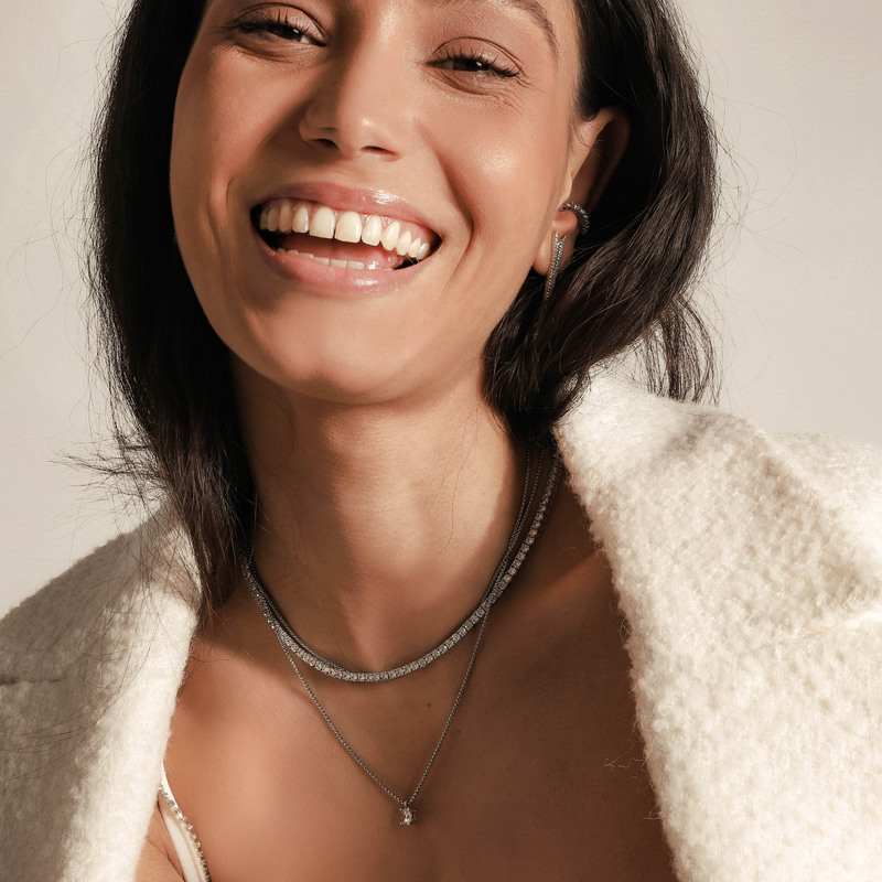 Model wearing the TENNIS NECKLACE SET which includes three layered necklaces. One is a 1mm wide, Stainless steel Rhodium plated chain, a Rhodium-plated brass/cubic zirconia tennis necklace and 18" necklace with a Solitaire 1 ct zirconia pendant. 
