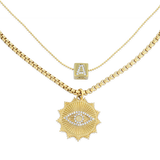The I See You Necklace, a layered gold necklace featuring two distinct chains. The shorter chain displays a delicate charm with an initial 'A' adorned with crystals, while the longer chain holds a bold sunburst pendant with an eye motif, encrusted with sparkling crystals. This layered design adds a mystical and personalized touch, perfect for making a unique statement