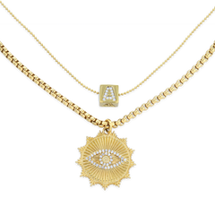 The I See You Necklace, a layered gold necklace featuring two distinct chains. The shorter chain displays a delicate charm with an initial 'A' adorned with crystals, while the longer chain holds a bold sunburst pendant with an eye motif, encrusted with sparkling crystals. This layered design adds a mystical and personalized touch, perfect for making a unique statement