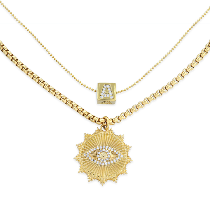 The I See You Necklace, a layered gold necklace featuring two distinct chains. The shorter chain displays a delicate charm with an initial 'A' adorned with crystals, while the longer chain holds a bold sunburst pendant with an eye motif, encrusted with sparkling crystals. This layered design adds a mystical and personalized touch, perfect for making a unique statement