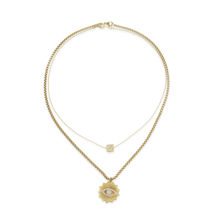 The I See You Necklace, a layered gold necklace featuring two chains. The shorter chain has a dainty square charm with an initial, while the longer chain showcases a striking sunburst pendant with an eye design encrusted in crystals. This mystical-inspired piece adds depth and personality, perfect for layering with other gold accessories or wearing solo for a bold look.