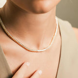 The Gold Tennis Necklace, a single gold necklace elegantly draped along the neckline, featuring a continuous row of sparkling round crystals. This timeless piece adds a sophisticated shimmer, perfect for both everyday elegance and special occasions.