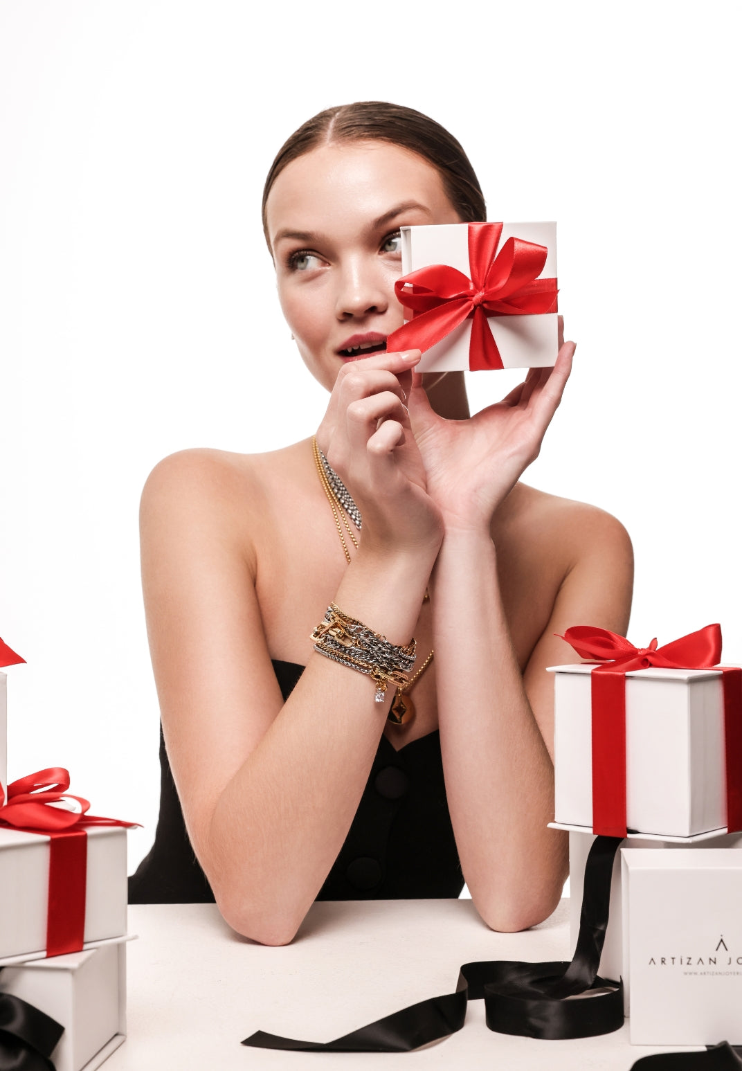 Woman holding an Artizan Joyería gift box with a red ribbon, styled with layered gold and silver necklaces and stacked bracelets featuring mixed metals and charms, surrounded by more gift boxes