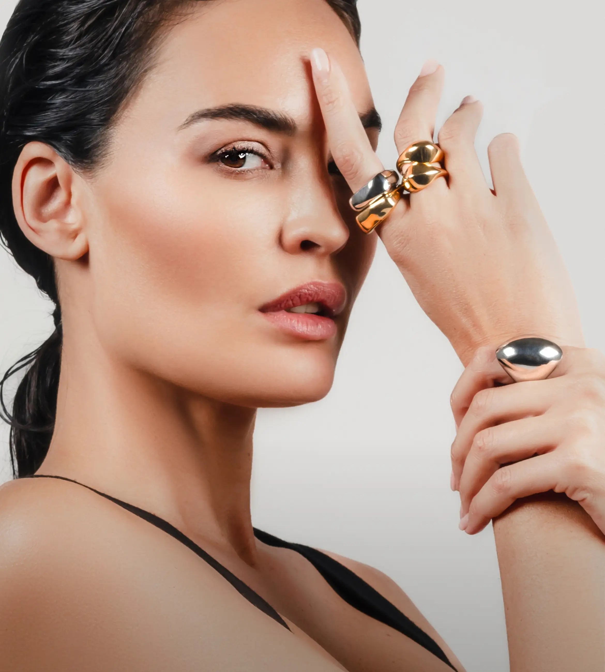 Close-up of a woman wearing bold mixed metal rings, including a gold and silver knot ring and a silver dome ring, with one hand resting on her forehead.