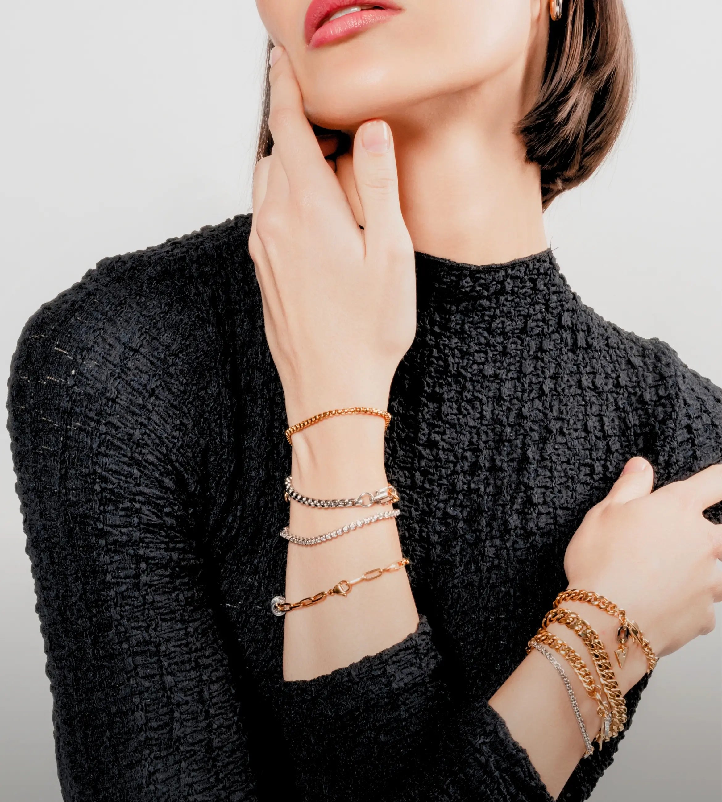 Close-up of a woman wearing a stack of mixed metal bracelets (gold and silver chains) and a black textured sweater, with one hand touching her neck.