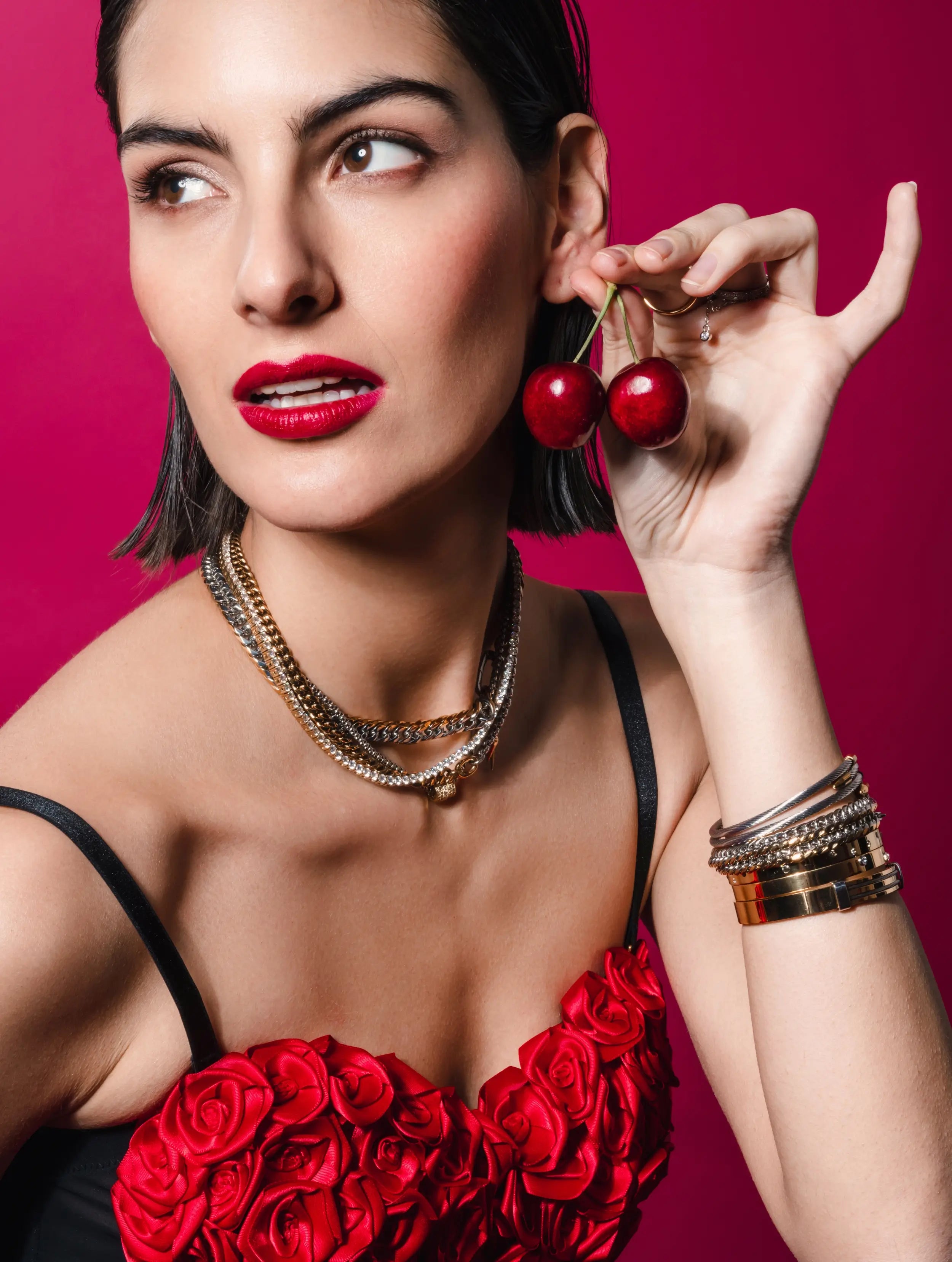 The image features a model holding cherries to her ear, wearing layered gold and silver necklaces and stacked bracelets. 