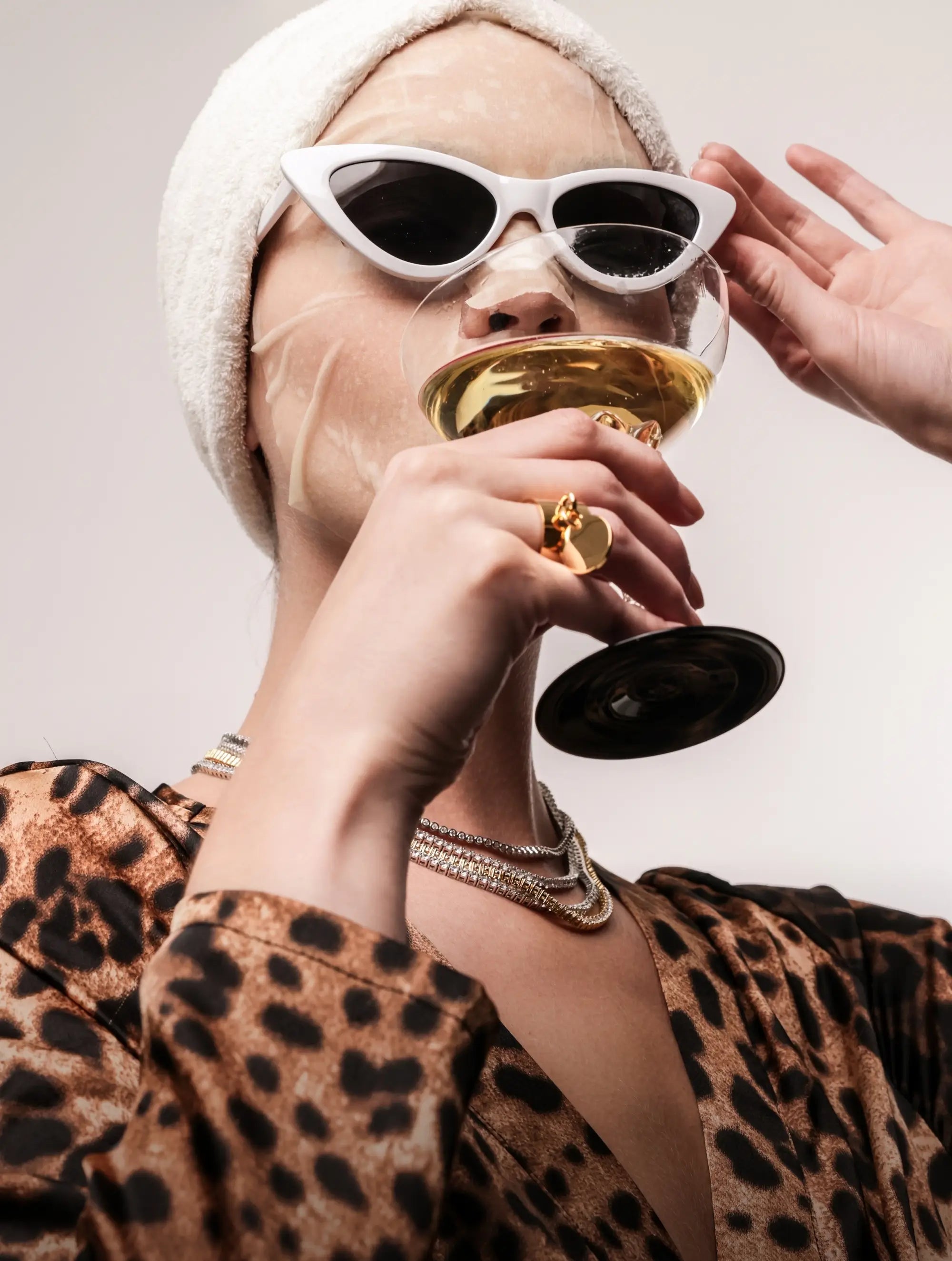 Woman in leopard robe and face mask sipping champagne, wearing three layered gold and silver necklaces and a chunky gold ring with charm details.