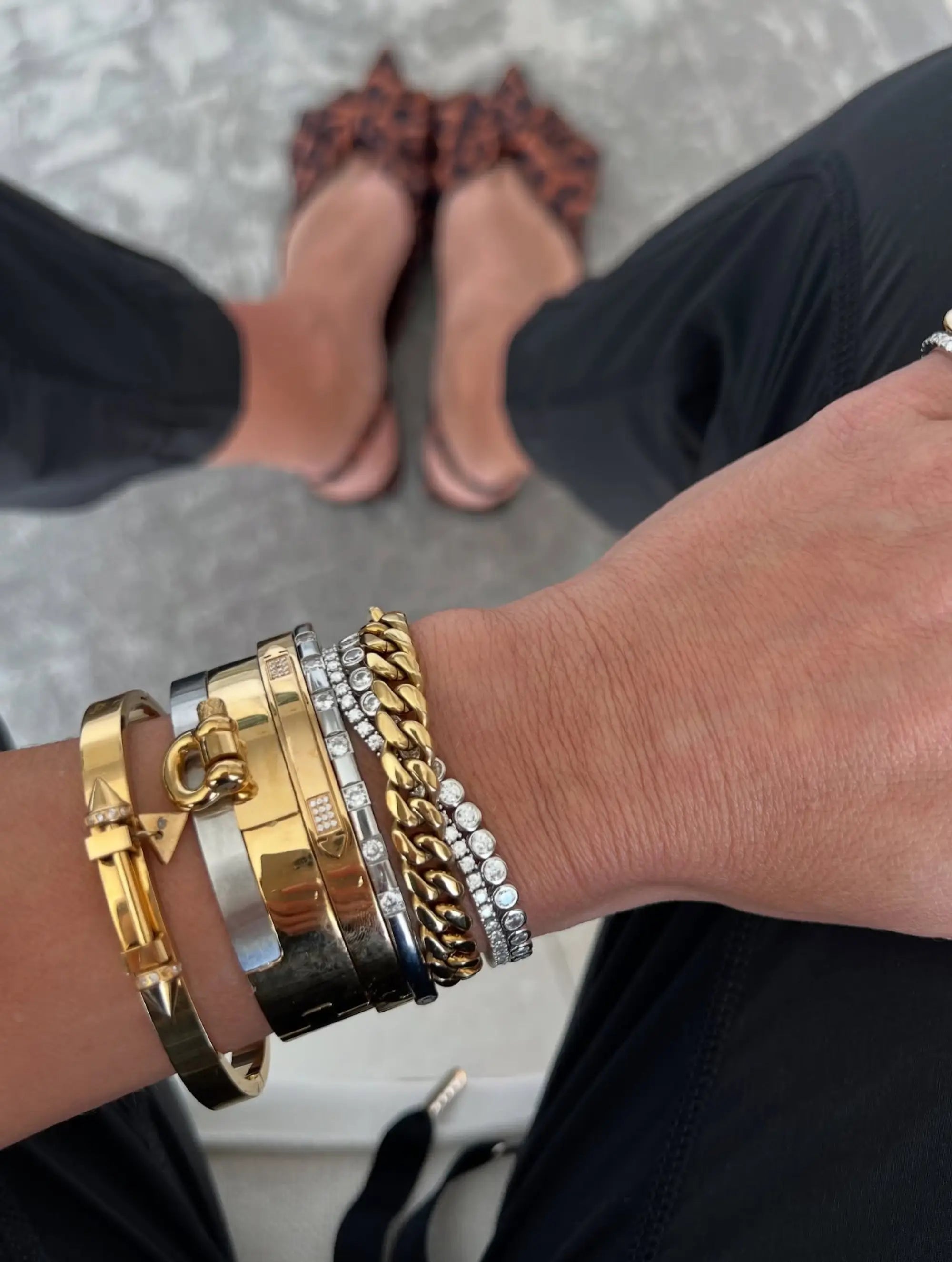 Woman's wrist featuring a bracelet stack with gold, silver, and diamond-like details, including a chunky chain bracelet, worn with black pants and leopard print shoes.