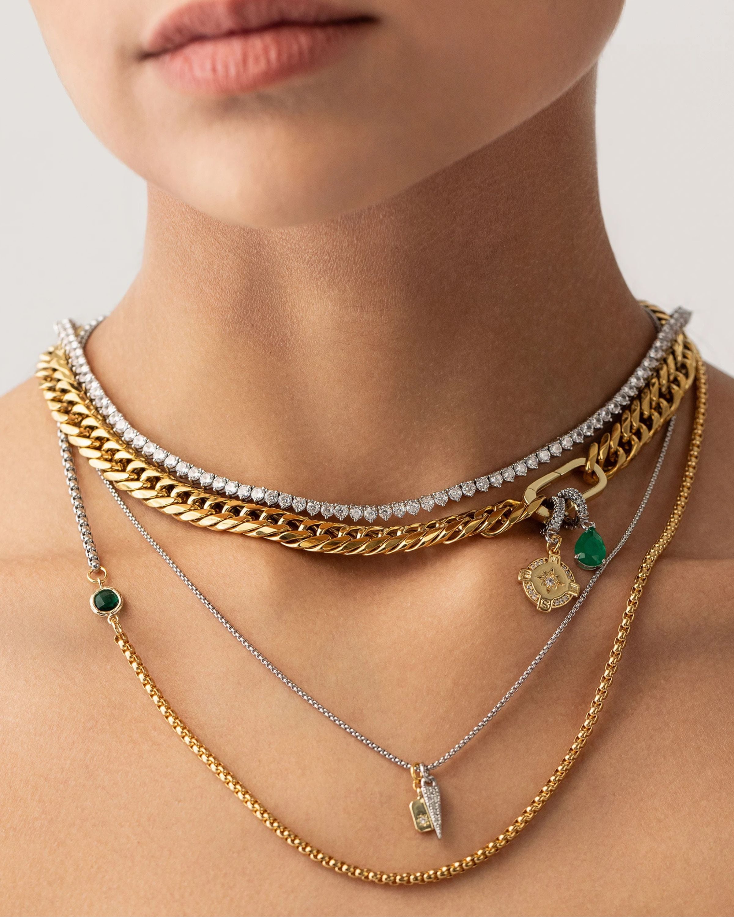 Model wearing The Just Click Emerald Tennis Necklace Set, a four-piece gold and silver necklace set featuring a variety of chains with diamond-like accents, emerald accents, and a variety of charms.