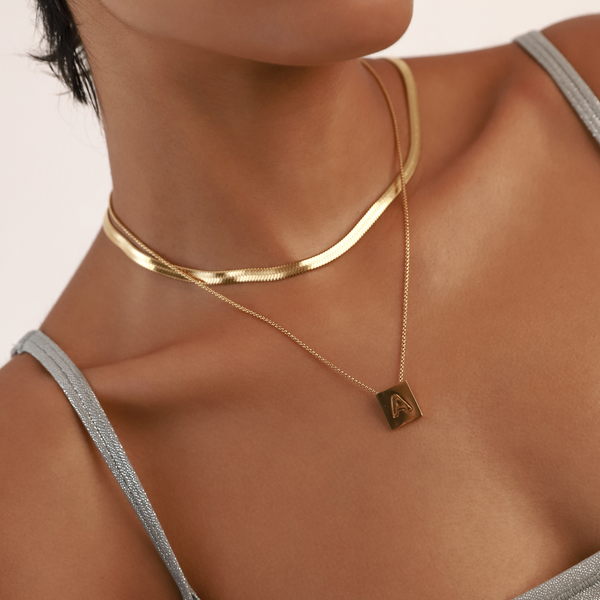 Model wearing the LETTER SQUARE NECKLACE made of Stainless steel chain 1mm wide chain and 18k gold plated square initial charm and a gold snake necklace. ||| MIX / A