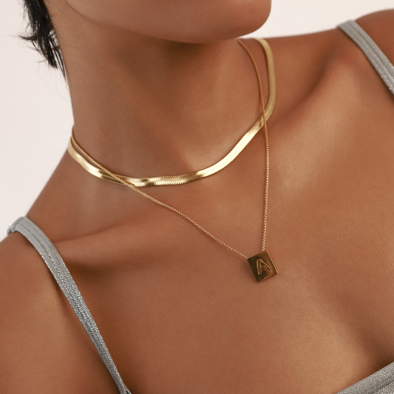 Model wearing the LETTER SQUARE NECKLACE made of Stainless steel chain 1mm wide chain and 18k gold plated square initial charm and a gold snake necklace. 