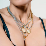 Woman wearing 5 layered necklaces in gold and silver with cross, heart, and medallion charms, styled over a dark green top. |||