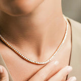 The Gold Tennis Necklace, a single gold necklace elegantly draped along the neckline, featuring a continuous row of sparkling round crystals. This timeless piece adds a sophisticated shimmer, perfect for both everyday elegance and special occasions.