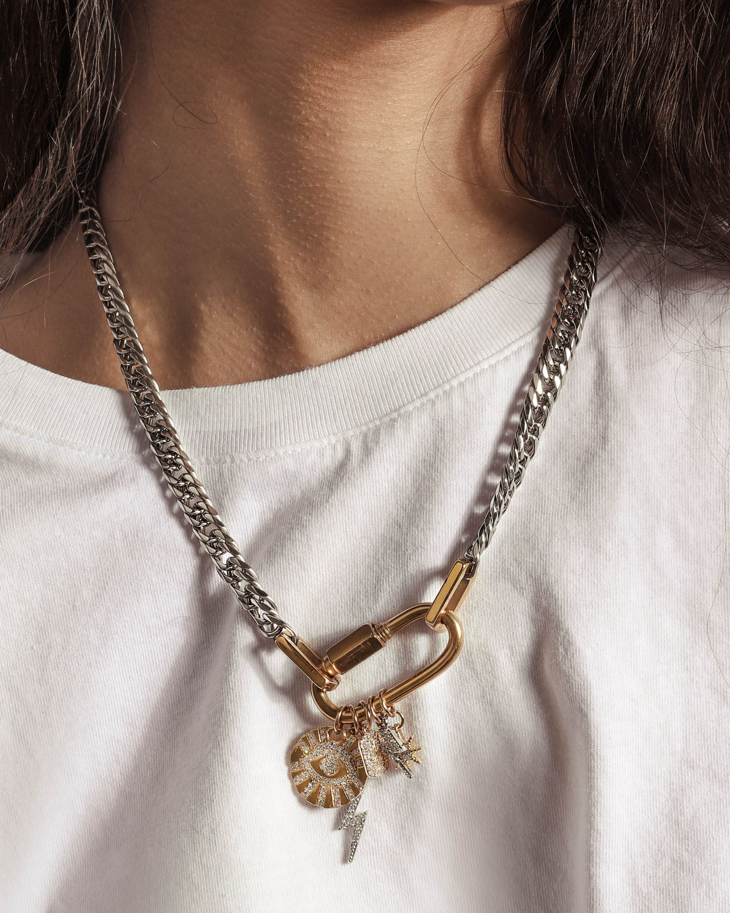 Model in a white shirt wearing the ANNE CHARM NECKLACE which is an 18", 9mm wide stainless steel chain with an 18K Gold Plated Stainless Steel carabiner clasp strung with dainty gold filled charms, including 2 lightning bolts, evil eye, celestial tag star and a star.