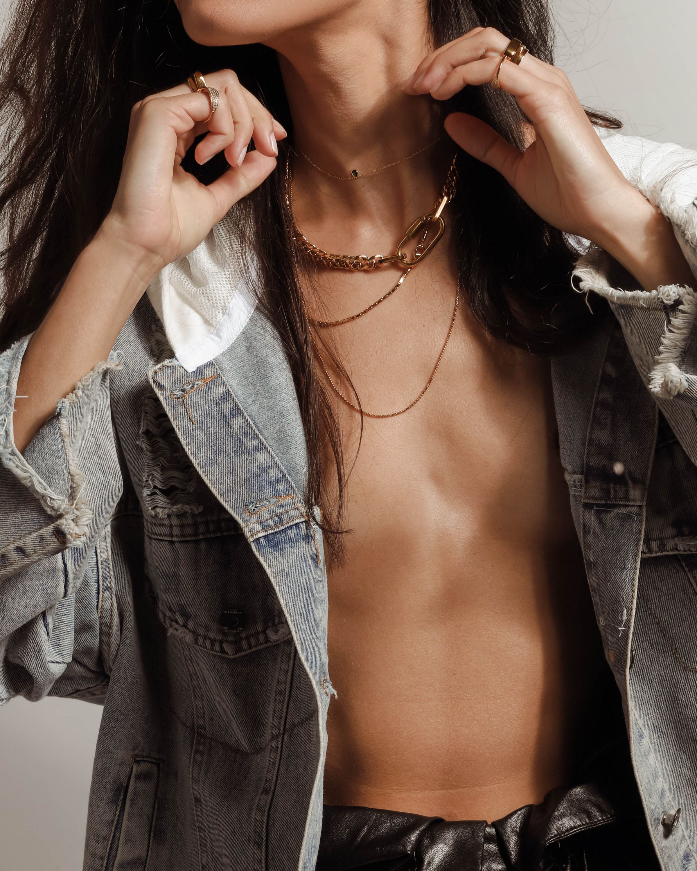 Model wearing an oversized denim jacket and The ANNE GOLD NECKLACE SET which includes 4 separate layered chains. The shortest is the Anne Necklace made of chunky 18k gold plated stainless steel chain with carabiner clasp followed with two thin necklaces that are 20" and 24" in length and a necklace with 925 Sterling Silver Gold plated emerald drop dainty charm.