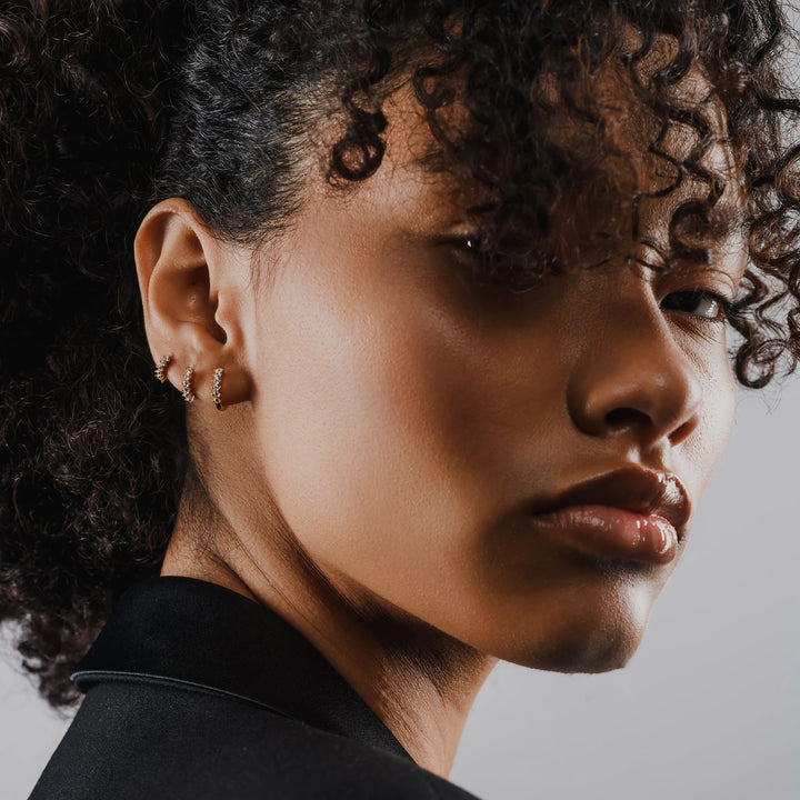 Model wearing 3 Tiny Huggies Hoop that are small gold hoop earrings that feature a row of sparkling cubic zirconia stones set in a  pointed, triangular pattern.||| GOLD