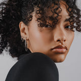 Model wearing the Endless Diamond Earring Chain, a gold chain featuring seven bezel-set diamond-like stones. She is also wearing two separate drop diamond earrings.