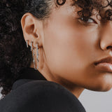 Model wearing the Endless Diamond Earring Chain, a gold chain featuring seven bezel-set diamond-like stones. She is also wearing two separate drop diamond earrings.