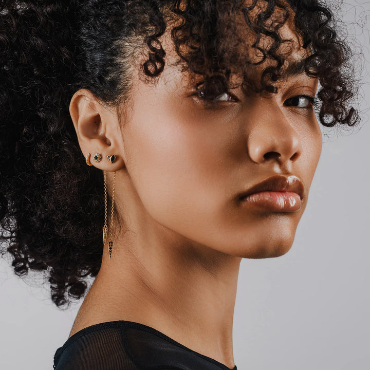 Model wearing the Arrow Earring Chain, a single gold chain earring with a V-shaped arrowhead pendant adorned with sparkling stones and 2 stud earrings.||| GOLD