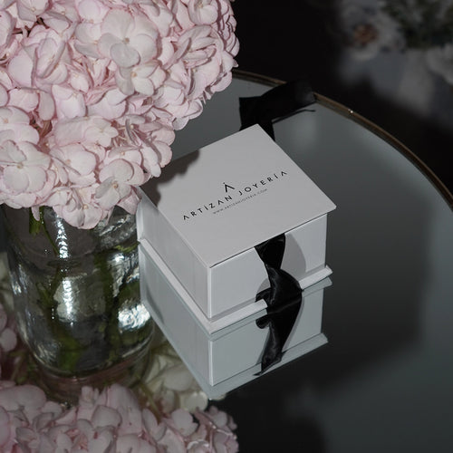 Elegant Artizan Joyeria gift box tied with a black ribbon, placed on a reflective surface beside a bouquet of soft pink flowers. The setup exudes a luxurious and sophisticated vibe, highlighting the brand's refined presentation.