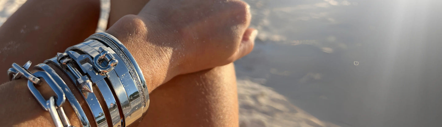 Close-up of stacked silver bracelets with bold links and charm details on a sandy wrist, photographed at the beach in warm sunset lighting.