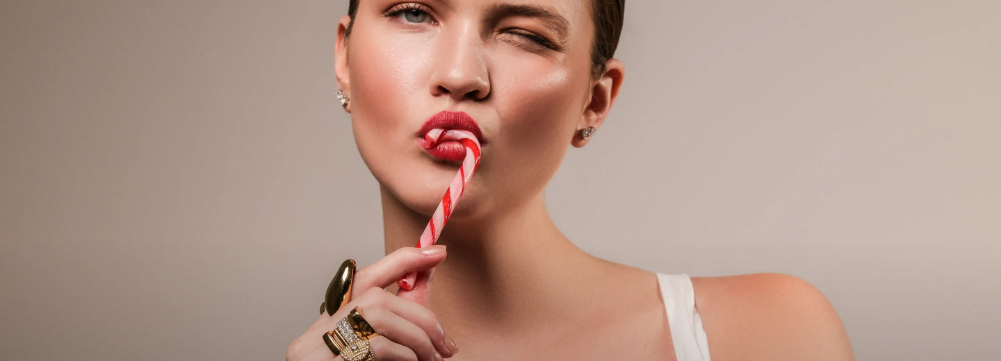 Close-up image of a model holding a red and white candy cane with her lips, wearing a white tank top and a bold stack of 5 gold statement rings, including crystal details and a lion-shaped design.