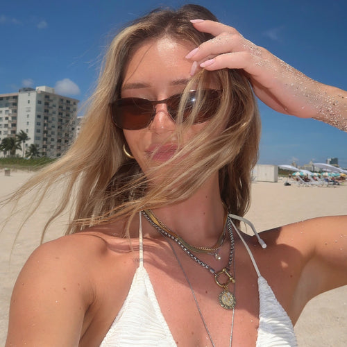 A woman at the beach with sandy hair and sunglasses, showcasing layered necklaces in gold and silver, complemented by a radiant smile under the sun.