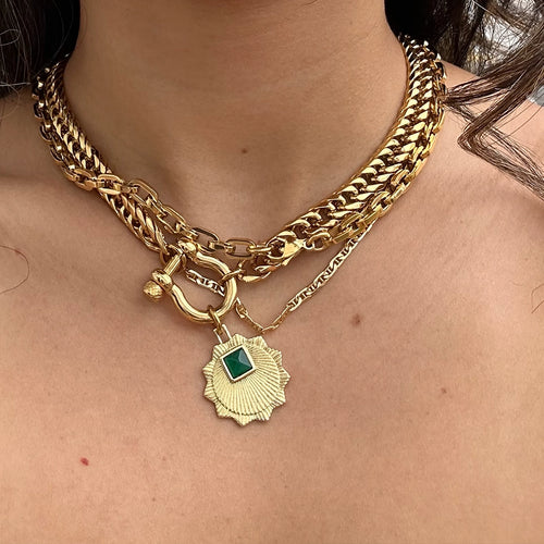 A bold gold chain necklace featuring a chunky link design and a prominent green gemstone pendant at the center, styled for a statement look. The necklace is layered against a simple, neutral background, emphasizing the rich contrast between the gold and the emerald stone.