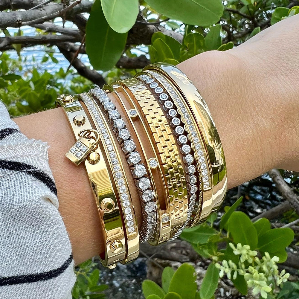 A woman's wrist adorned with an Artizan stack of gold and diamond bracelets, including a bangle with a padlock charm, a diamond-studded woven bracelet, and several other gold bangles.