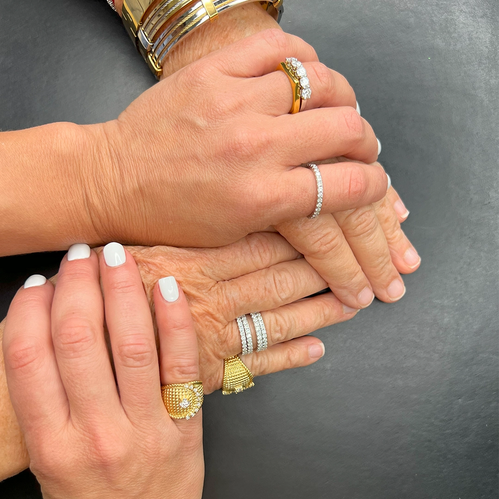 Close-up of three hands stacked together, each adorned with a variety of gold and silver rings with intricate designs and diamond accents. The image showcases generational style and elegance, highlighting the timeless appeal of mixed-metal jewelry.