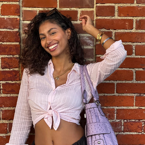 A smiling woman in a light pink blouse with Artizan Joyeria necklaces and bracelets, paired with a pastel lavender bag against a brick wall