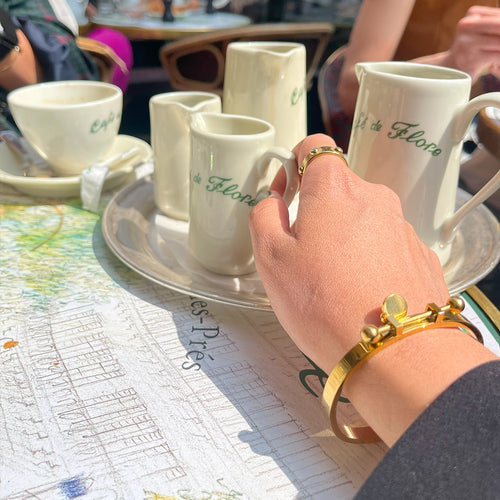 Hand adorned with gold rings and a gold bangle resting on a classic coffee shop table with cups and pitchers.