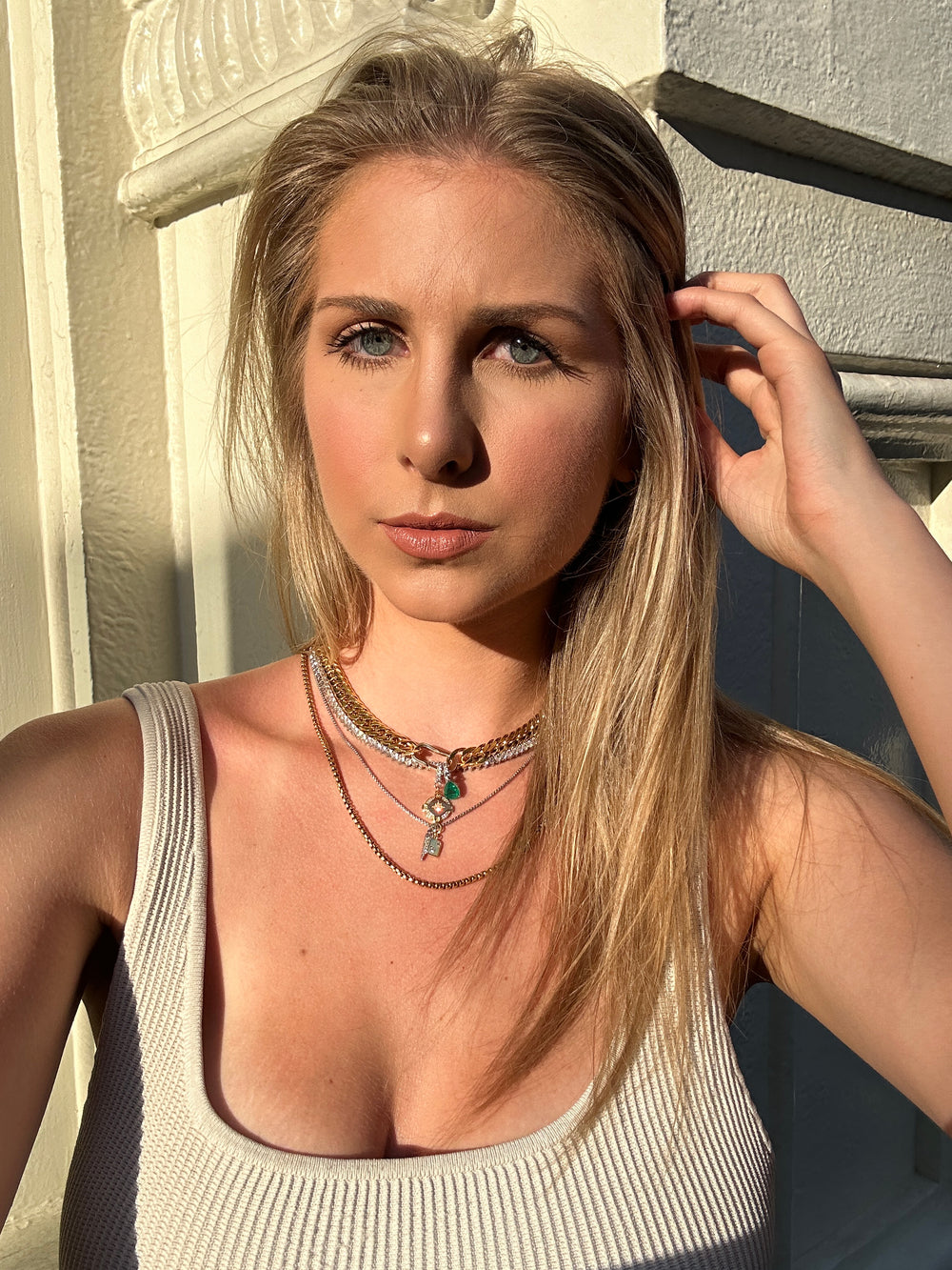 Model wearing layered silver and gold necklaces with a green stone charm, posing outdoors with soft lighting. She is dressed in a white tank top with natural makeup and long blonde hair.