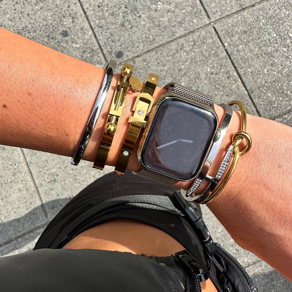 A woman's wrist wearing a stack of gold and silver bracelets, a silver cuff bracelet with a bold design all of these from artizan joyeria, and an Apple Watch with a Milanese mesh band.