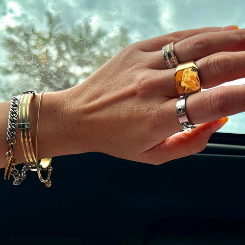 Hand with layered bracelets and rings, including mixed metals and gold accents, with a scenic view in the background.