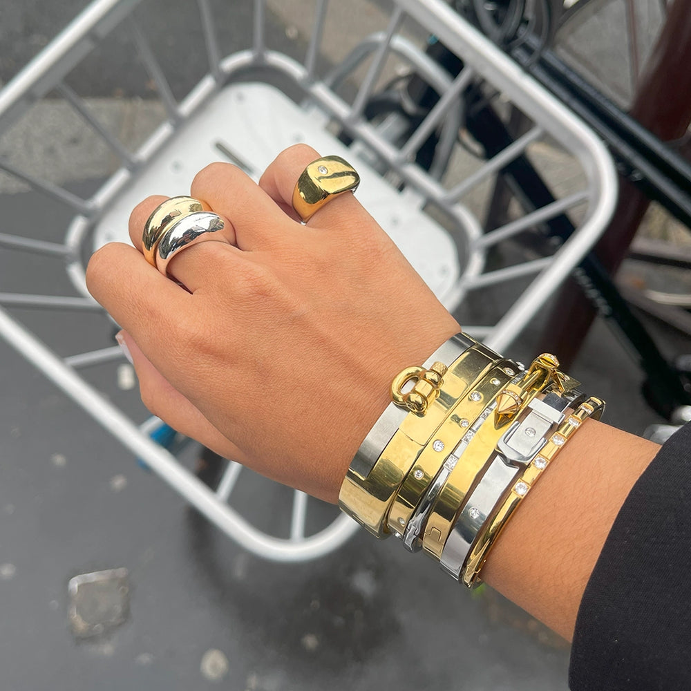 Close-up of a hand adorned with stacked gold and silver rings and bracelets, set against a backdrop of a metal basket. The mixed-metal accessories create a bold, chic look