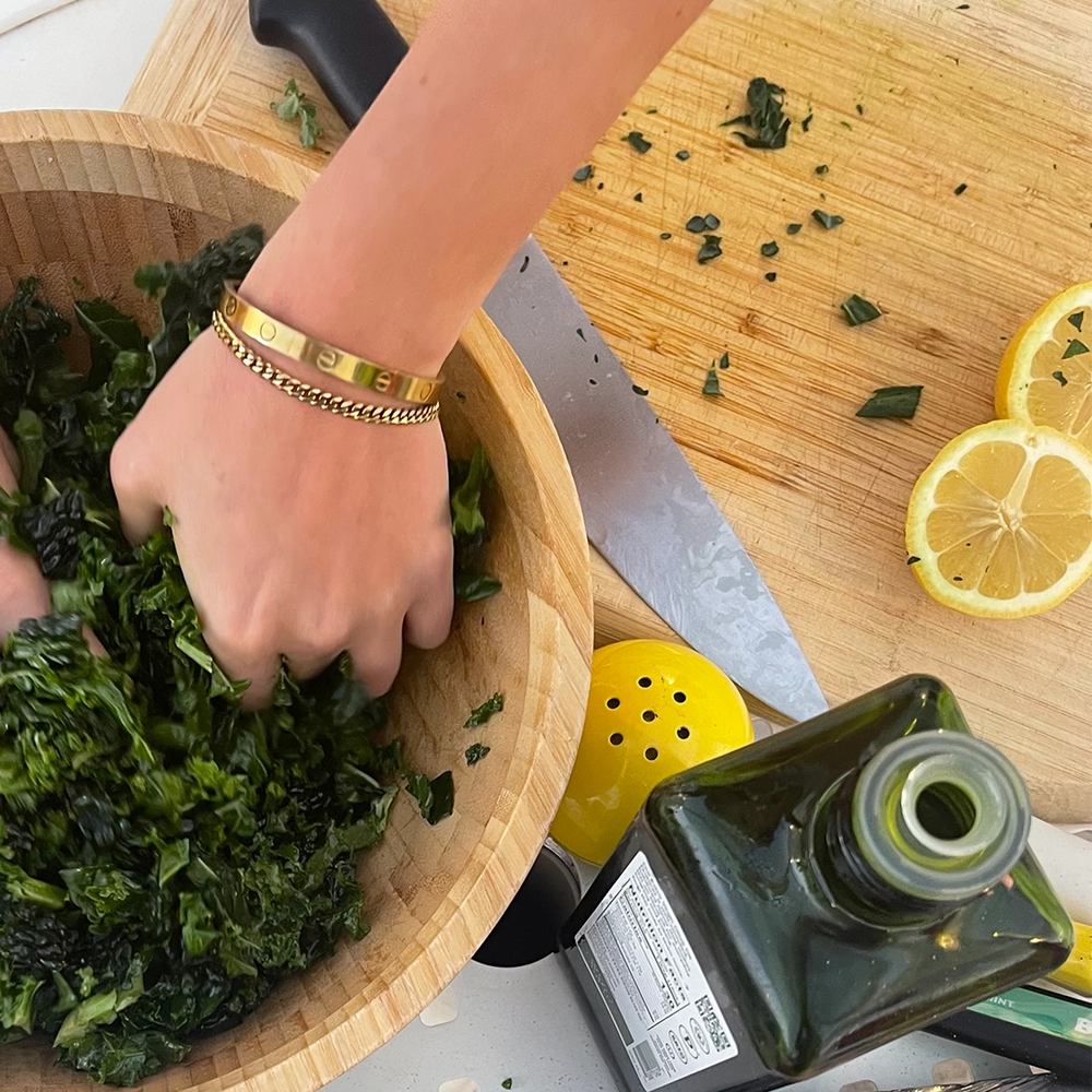 Hands mixing salad in a wooden bowl, showcasing gold bracelets and statement rings. The image combines fashion with a healthy, casual lifestyle.