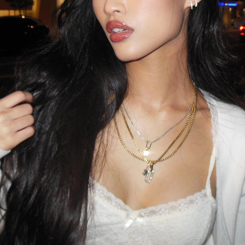 A close-up of a model wearing a gold necklace with a pendant, elegantly styled with a white lace camisole and soft lighting accentuating her features