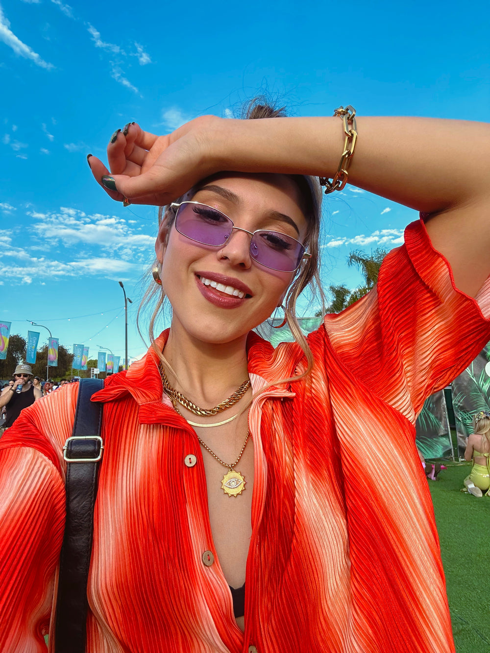 A woman wearing a red and orange striped shirt, layered gold necklaces including an evil eye pendant, a chunky chain necklace, and a bracelet. She is smiling, with sunglasses and a backdrop of blue sky and palm trees.