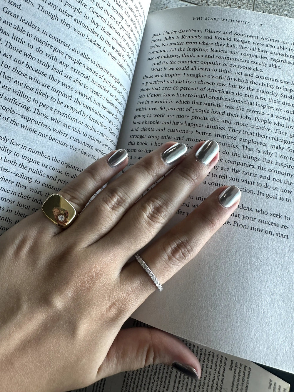 A close-up of two hands with metallic silver nails, showcasing a gold ring with a heart design and a delicate silver band, resting on an open book with visible text.