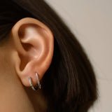 The MINI SHOT HOOP, which is a pair of elegant silver earrings, showcases a minimalist hoop design encrusted with shimmering cubic zirconia stones and finished with a sleek pointed charm for a modern and edgy touch. Perfect for enhancing any outfit with a subtle yet sophisticated sparkle.