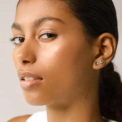 The Emerald Flower Stud Earring, shown as part of an ear stack with three other gold and black gemstone earrings. This dainty gold stud features a green emerald at the center, surrounded by a halo of sparkling stones in a floral design, adding an elegant and unique touch to a layered ear look.