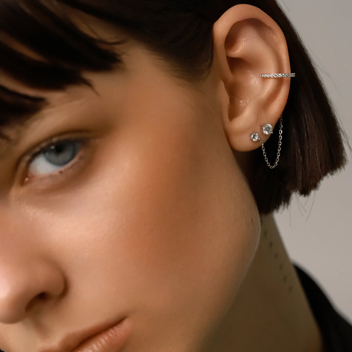 The Ear Cuff with Chain Set, which is a four-pieceThe Ear Cuff with Chain Set, which is a four-piece silver earring set, features a sparkling ear cuff connected by a delicate chain to a stud, accompanied by two additional solitaire studs for versatile styling options and an elegant, contemporary look.
