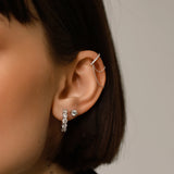 The Main Hoop, which is a pair of silver-tone hoop earrings adorned with a row of sparkling round-cut stones, showcased here alongside a delicate stud earring and ear chain for a layered, sophisticated ear stack look