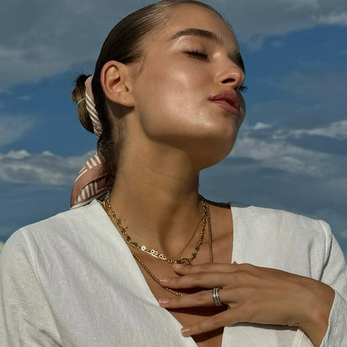 A close-up of a woman wearing layered gold necklaces with delicate chains and small pendants, styled against a clear blue sky. The gold necklaces include a thin curb chain and a pendant necklace, creating an elegant, everyday look perfect for sunny weather.