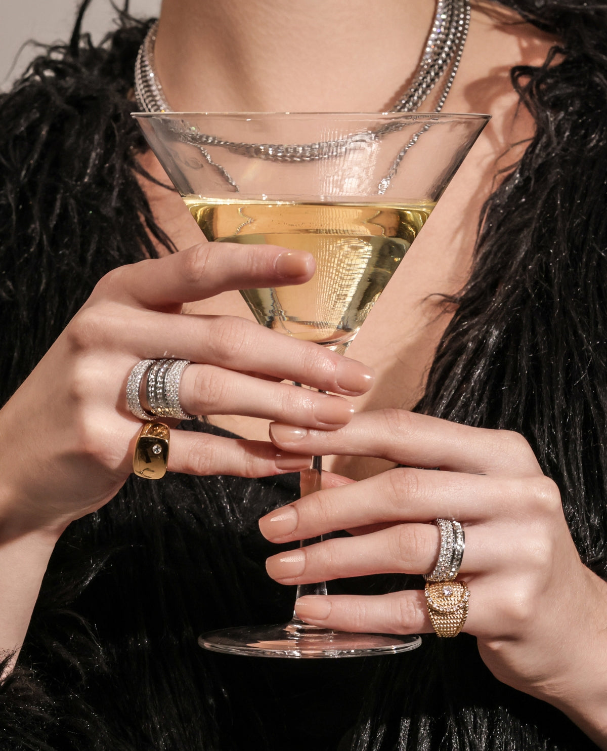 Close-up of a woman's hands holding a martini glass, adorned with multiple gold and silver rings that add a glamorous touch. She wears layered necklaces and a chic black outfit, exuding an elegant and sophisticated vibe.