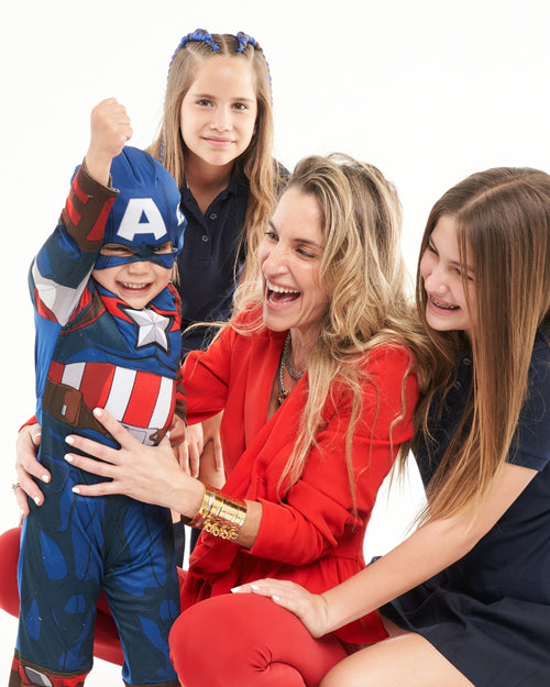 A vibrant mother in a red outfit, laughing with her three children, one of whom is dressed as Captain America. She wears bold gold jewelry, showcasing how stylish accessories elevate everyday moments.