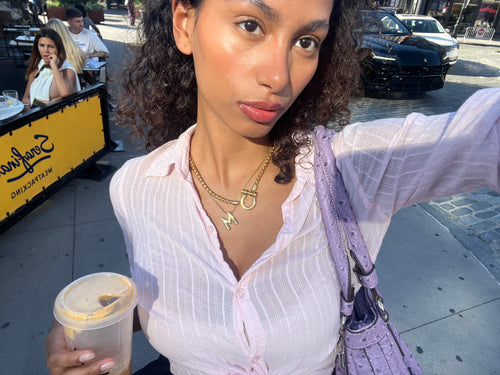 A woman in a light pink blouse, accessorized with Artizan Joyeria’s layered necklaces and bracelets, holding an iced coffee on a sunny city street