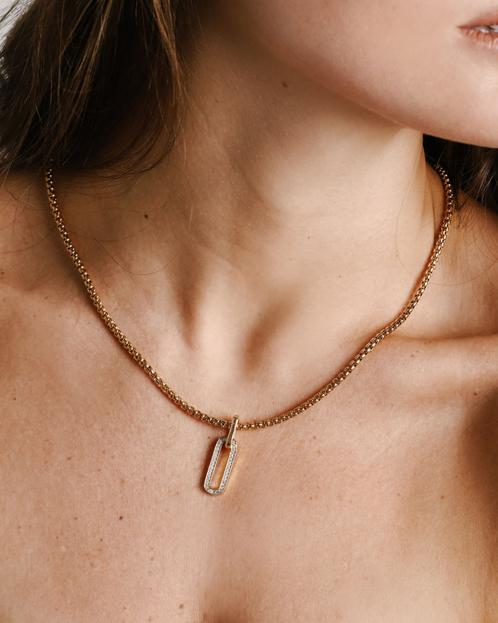 Model is wearing the PROCIDA NECKLACE which is a 20" Stainless steel 18k gold plated chain with an oval shaped gold plated and Zirconia charm.