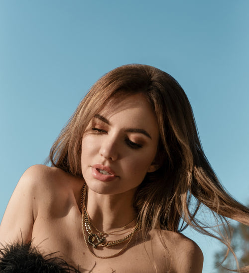 A woman with soft waves in her brown hair poses gracefully against a bright blue sky background. She wears layered gold necklaces, including a statement chain with a central decorative piece, offering a sophisticated yet contemporary look. The sunlight accentuates her relaxed and natural pose, enhancing the warm tones of the jewelry.